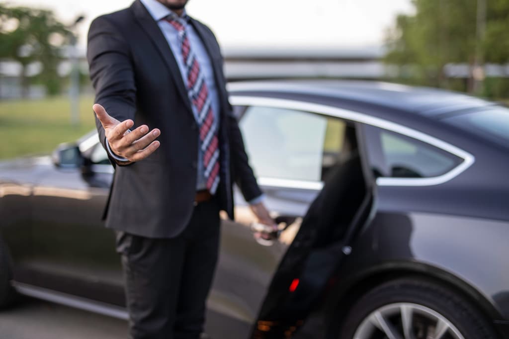 A chauffeur in a suit gestures a passenger toward the open door of a dark sedan