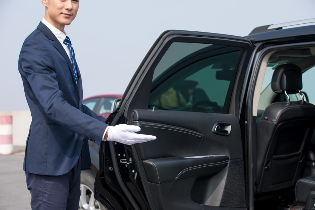 A smiling chauffeur in white gloves holds open the rear door of a black SUV